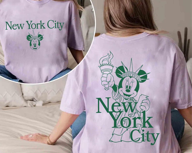 May include: A light purple t-shirt with the text "New York City" in green, along with a green graphic of Minnie Mouse as the Statue of Liberty. The back of the shirt also features the same design.