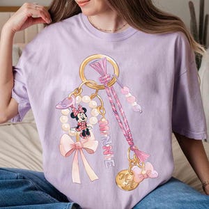 Op de afbeelding: Een lichtpaars T-shirt met een Minnie Mouse sleutelhanger ontwerp. De sleutelhanger bevat een gouden ring, parels, strikken en de naam "Minnie" in roze. Het ontwerp is een grillige en kleurrijke illustratie.