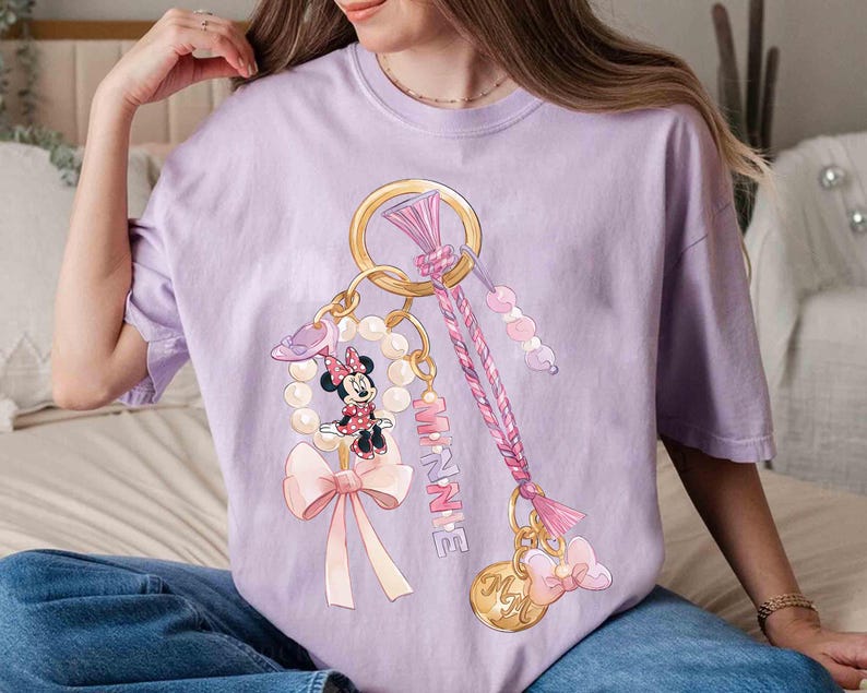 Op de afbeelding: Een lichtpaars T-shirt met een Minnie Mouse sleutelhanger ontwerp. De sleutelhanger bevat een gouden ring, parels, strikken en de naam "Minnie" in roze. Het ontwerp is een grillige en kleurrijke illustratie.
