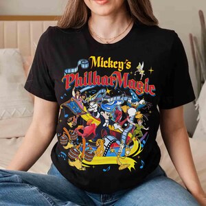 K&ouml;nnte beinhalten: Schwarzes T-Shirt mit einer farbenfrohen Grafik von Mickey Mouse und anderen Disney-Figuren. Der Text "Mickey's PhilharMagic" ist in Gelb, Rot und Blau gehalten. Das Design zeigt Aladdin, Peter Pan und Simba.