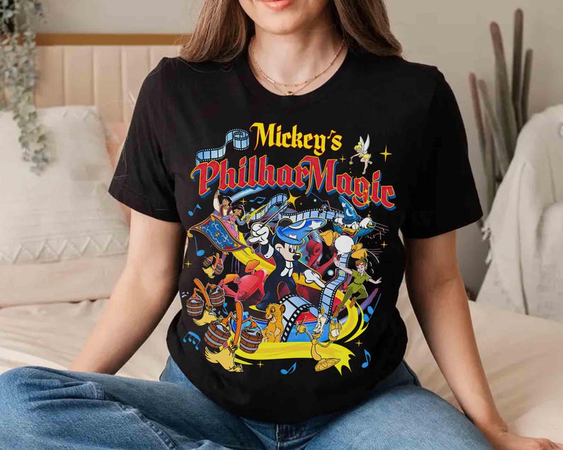 K&ouml;nnte beinhalten: Schwarzes T-Shirt mit einer farbenfrohen Grafik von Mickey Mouse und anderen Disney-Figuren. Der Text "Mickey's PhilharMagic" ist in Gelb, Rot und Blau gehalten. Das Design zeigt Aladdin, Peter Pan und Simba.
