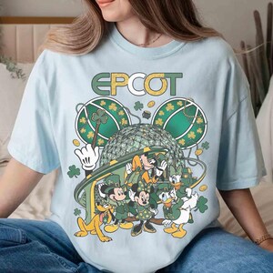 May include: Light blue t-shirt featuring a graphic of Disney characters celebrating St. Patrick's Day at Epcot. The design includes Mickey, Minnie, Donald, Daisy, Goofy, and Pluto, with shamrocks and the Epcot logo.