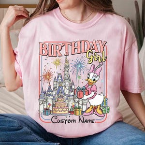 May include: Pink t-shirt with a graphic featuring Daisy Duck holding a gift in front of a castle, fireworks, and a birthday cake. Text reads "BIRTHDAY Girl" and "Custom Name".