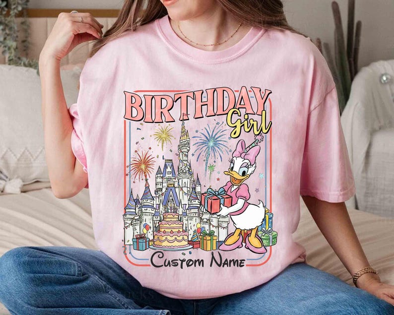 May include: Pink t-shirt with a graphic featuring Daisy Duck holding a gift in front of a castle, fireworks, and a birthday cake. Text reads "BIRTHDAY Girl" and "Custom Name".