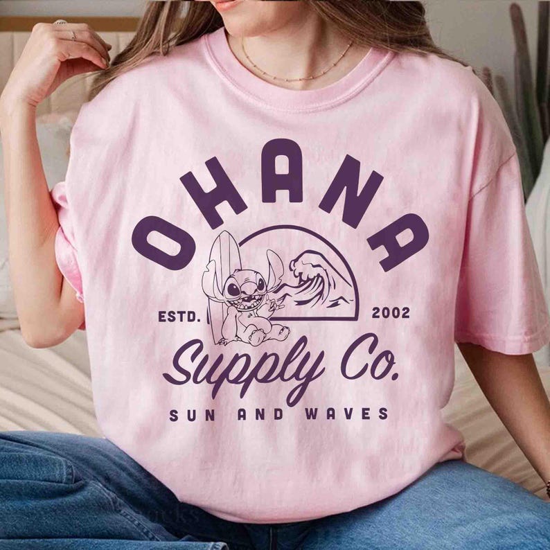 May include: Pink t-shirt with a purple graphic featuring the word "OHANA" above a cartoon character with a surfboard and a wave. The text "Supply Co." and "Sun and Waves" are also included.