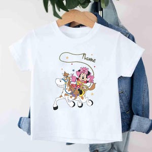 May include: White t-shirt featuring a cartoon Minnie Mouse riding a horse with a lasso. Minnie Mouse is wearing a pink cowgirl outfit and a matching bow. The word "Name" is written in cursive above the graphic.