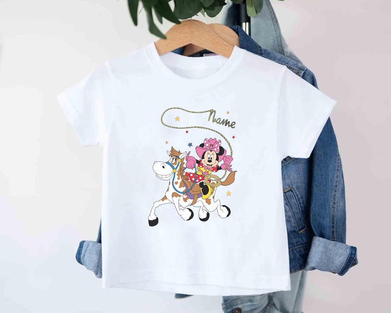 May include: White t-shirt featuring a cartoon Minnie Mouse riding a horse with a lasso. Minnie Mouse is wearing a pink cowgirl outfit and a matching bow. The word "Name" is written in cursive above the graphic.