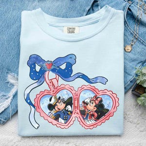 May include: Light blue t-shirt with a print of Mickey Mouse and Minnie Mouse in heart-shaped frames. The frame is decorated with a blue bow and a ribbon. The text "COMFORT COLORS" is printed on the shirt.