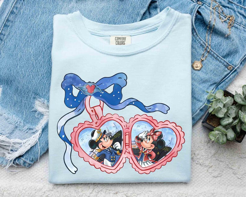 May include: Light blue t-shirt with a print of Mickey Mouse and Minnie Mouse in heart-shaped frames. The frame is decorated with a blue bow and a ribbon. The text "COMFORT COLORS" is printed on the shirt.