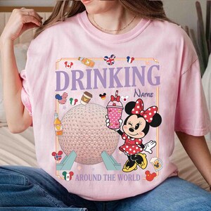 Puede incluir: Camiseta rosa claro con un dise&ntilde;o de Minnie Mouse sosteniendo una bebida, el texto "DRINKING" y "AROUND THE WORLD". El dise&ntilde;o incluye un globo terr&aacute;queo, iconos de pa&iacute;ses y un campo de nombre.