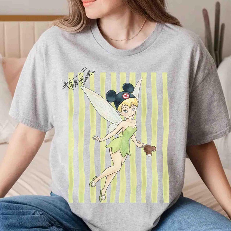 May include: A heather gray t-shirt featuring a graphic of Tinker Bell wearing black Mickey Mouse ears. The design includes vertical yellow stripes in the background and the handwritten text Tinker Bell in black ink above the character.