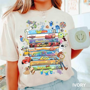 May include: Ivory-colored t-shirt featuring a stack of colorful Disney movie covers, including "Toy Story," "Cars," and "Monsters, Inc." Various animated characters surround the movie titles. A white mug with a smiley face is held in the image.