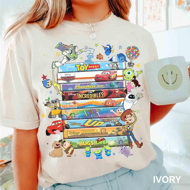 May include: Ivory-colored t-shirt featuring a stack of colorful Disney movie covers, including "Toy Story," "Cars," and "Monsters, Inc." Various animated characters surround the movie titles. A white mug with a smiley face is held in the image.