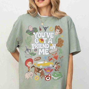 May include: Sage green t-shirt featuring the phrase "You've Got a Friend in Me" with cartoon character illustrations. The design includes a cactus, a rocket, a horseshoe, and a cowboy boot.