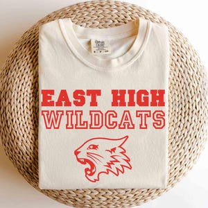 Può includere: Una maglietta bianca con testo rosso che recita "East High Wildcats" e un'immagine di un gatto selvatico di cartone rosso.