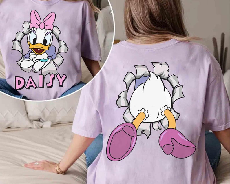 Puede incluir: Camiseta lila claro con un dise&ntilde;o de Daisy Duck. La parte delantera muestra a Daisy Duck saliendo de un agujero rasgado, con el nombre "DAISY" debajo. La parte trasera de la camiseta muestra la parte trasera y los pies de Daisy.