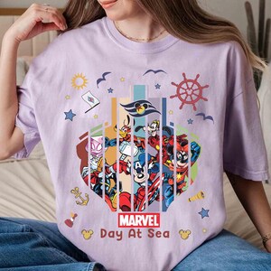 May include: A light purple t-shirt featuring a colorful graphic of Marvel characters and Disney characters. The graphic includes the text "MARVEL Day At Sea" and nautical elements like a ship's wheel, anchor, and stars.