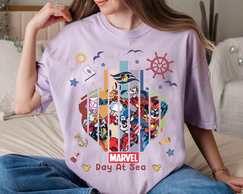 May include: A light purple t-shirt featuring a colorful graphic of Marvel characters and Disney characters. The graphic includes the text "MARVEL Day At Sea" and nautical elements like a ship's wheel, anchor, and stars.