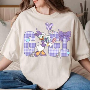 May include: Cream-colored t-shirt featuring a graphic of Daisy Duck holding a Mickey Mouse balloon. The letters "G" and "I" are in a purple plaid pattern, with floral and heart accents. The shirt is a relaxed fit.