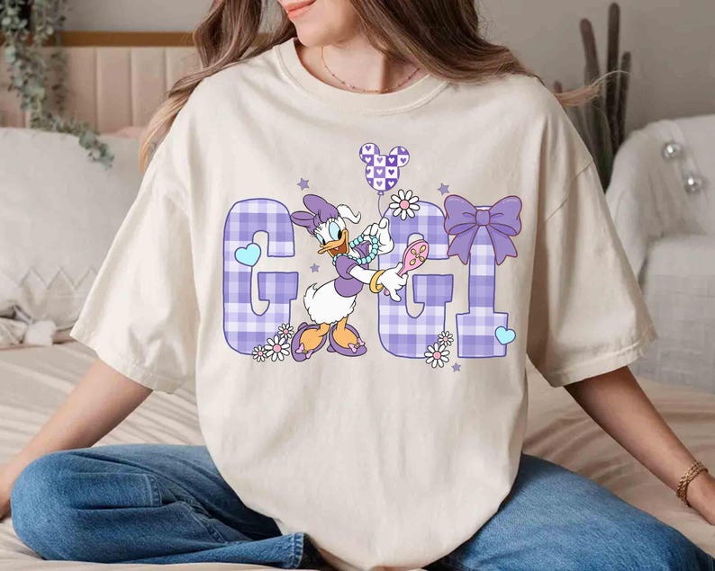 May include: Cream-colored t-shirt featuring a graphic of Daisy Duck holding a Mickey Mouse balloon. The letters "G" and "I" are in a purple plaid pattern, with floral and heart accents. The shirt is a relaxed fit.