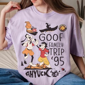 Pode incluir: Camiseta lil&aacute;s clara com um desenho do Pateta e Max do filme "Pateta e Max". O design inclui o texto "Goof Family Trip '95" e "Ahyuck!" com elementos de desenho animado.