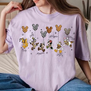May include: A light purple t-shirt featuring cartoon characters holding Mickey Mouse-shaped balloons. The characters include Pluto, Goofy, Mickey Mouse, Minnie Mouse, Donald Duck, and Daisy Duck. Each character's name is written below them.