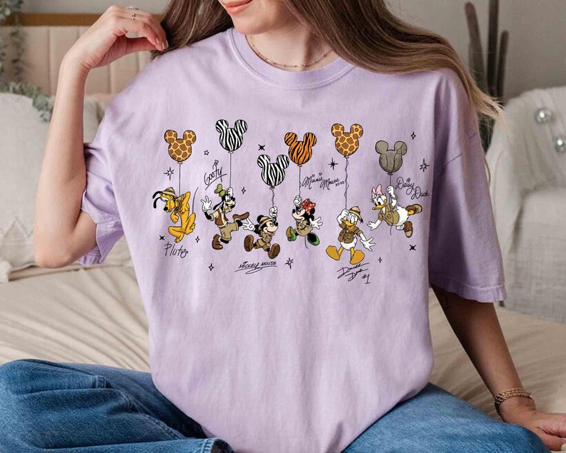 May include: A light purple t-shirt featuring cartoon characters holding Mickey Mouse-shaped balloons. The characters include Pluto, Goofy, Mickey Mouse, Minnie Mouse, Donald Duck, and Daisy Duck. Each character's name is written below them.