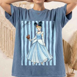 May include: A blue short-sleeve t-shirt featuring a graphic of Cinderella wearing a black mouse-ear headband and holding a chocolate-covered ice cream bar. The design includes vertical light blue stripes and the cursive text Cinderella.