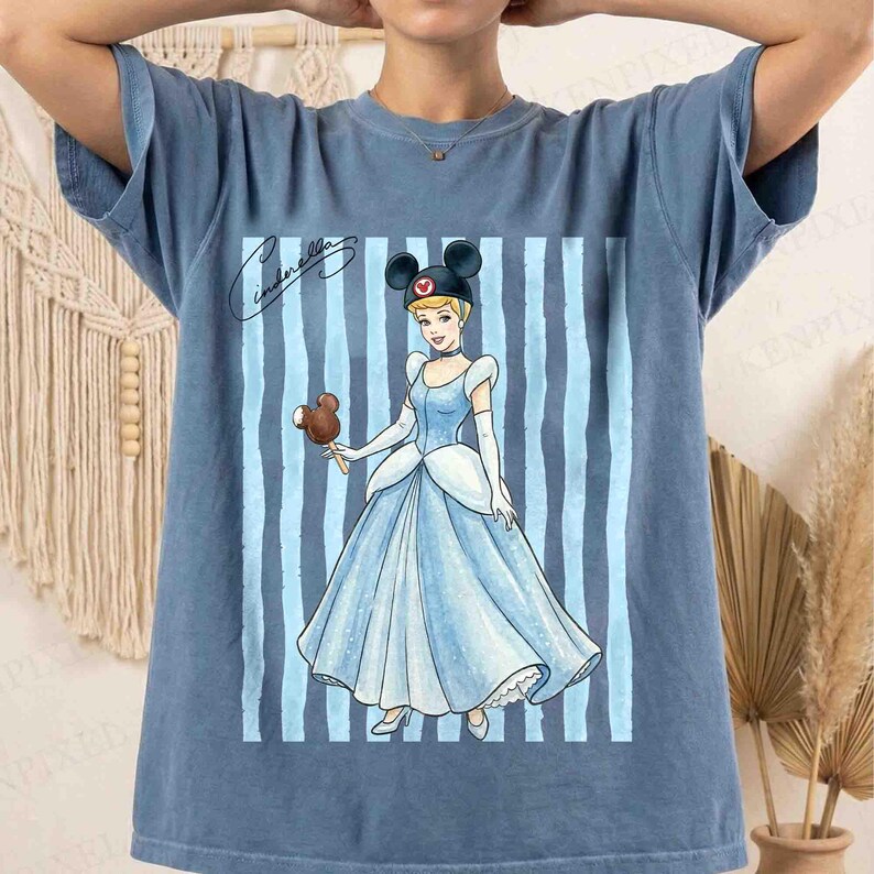 May include: A blue short-sleeve t-shirt featuring a graphic of Cinderella wearing a black mouse-ear headband and holding a chocolate-covered ice cream bar. The design includes vertical light blue stripes and the cursive text Cinderella.