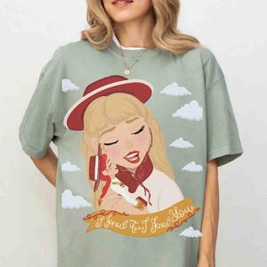 May include: A sage green short-sleeve t-shirt featuring a graphic illustration of a blonde character wearing a red hat, holding a small doll. The design includes white clouds and a tan banner with the text I Knew It, I Knew You.