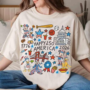 Puede incluir: Camiseta color crema con un gr&aacute;fico colorido que celebra el 250 aniversario de Am&eacute;rica. El dise&ntilde;o incluye personajes de dibujos animados, el Capitolio, un puente, estrellas y el texto "HAPPY 250 AMERICA 2026".