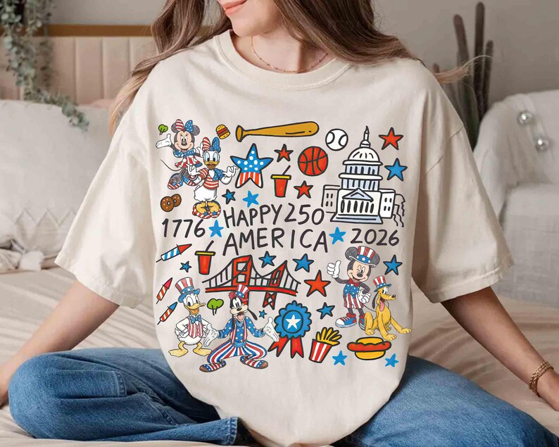 Puede incluir: Camiseta color crema con un gr&aacute;fico colorido que celebra el 250 aniversario de Am&eacute;rica. El dise&ntilde;o incluye personajes de dibujos animados, el Capitolio, un puente, estrellas y el texto "HAPPY 250 AMERICA 2026".