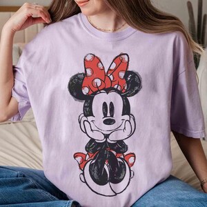 May include: A lilac t-shirt with a Minnie Mouse graphic. The cartoon character is in black and white with a red polka dot bow and shoes. The t-shirt has a loose fit.