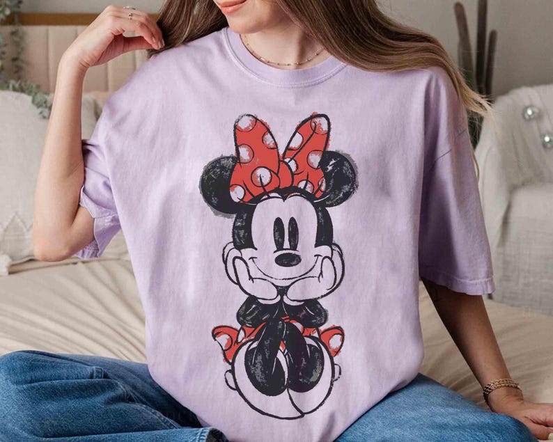 May include: A lilac t-shirt with a Minnie Mouse graphic. The cartoon character is in black and white with a red polka dot bow and shoes. The t-shirt has a loose fit.