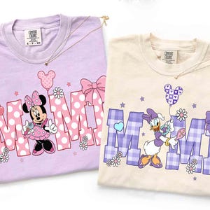 Puede incluir: Dos camisetas Comfort Colors, una lavanda y otra crema, con personajes de Disney. La camiseta lavanda tiene a Minnie Mouse con la palabra "MIMI" en un dise&ntilde;o de lunares. La camiseta crema tiene a Daisy Duck con la palabra "MIMI" en un dise&ntilde;o de cuadros.
