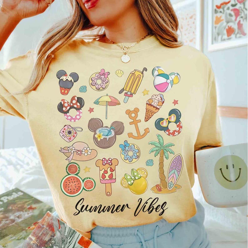 以下が含まれることがあります： 夏をテーマにした黄色のTシャツ。ミッキーマウス、アイスクリーム、スイカ、サーフボードなど、さまざまなカラフルな漫画の画像が描かれています。Tシャツには「Summer Vibes」という文字がプリントされています。