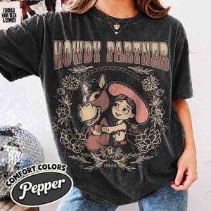 Puede incluir: Una camiseta de manga corta gris carb&oacute;n con el texto HOWDY PARTNER en una fuente degradada. El gr&aacute;fico incluye un caballo de dibujos animados y un personaje con sombrero, rodeados por una corona floral y la palabra PIXAR impresa abajo.