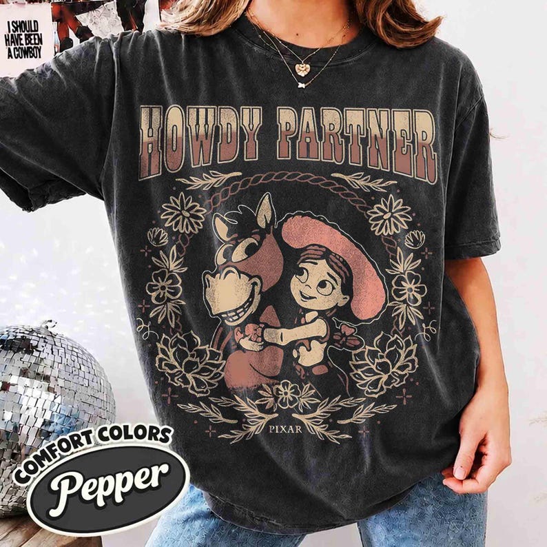 Puede incluir: Una camiseta de manga corta gris carb&oacute;n con el texto HOWDY PARTNER en una fuente degradada. El gr&aacute;fico incluye un caballo de dibujos animados y un personaje con sombrero, rodeados por una corona floral y la palabra PIXAR impresa abajo.