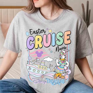 K&ouml;nnte beinhalten: Ein graues T-Shirt mit einem farbenfrohen Grafikdesign. Das Design enth&auml;lt den Text "Easter CRUISE Name", ein Cartoon-Bild von Daisy Duck in einem Osterhasen-Kost&uuml;m, ein Kreuzfahrtschiff und die Worte "Disney Treasure".