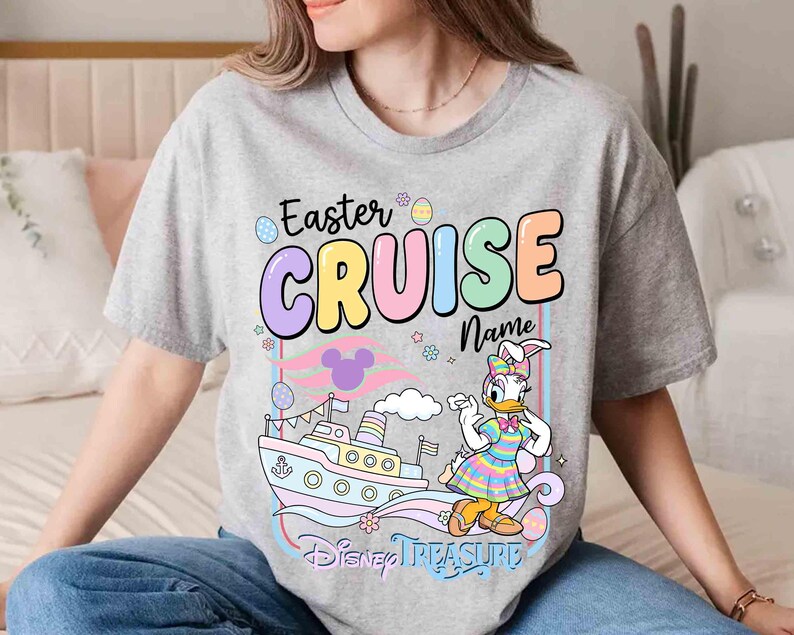 K&ouml;nnte beinhalten: Ein graues T-Shirt mit einem farbenfrohen Grafikdesign. Das Design enth&auml;lt den Text "Easter CRUISE Name", ein Cartoon-Bild von Daisy Duck in einem Osterhasen-Kost&uuml;m, ein Kreuzfahrtschiff und die Worte "Disney Treasure".