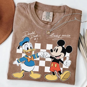 May include: Brown t-shirt featuring Donald Duck and Mickey Mouse fist-bumping, with the text "Donald Duck #1" and "Mickey Mouse". The shirt has a checkerboard pattern and a Comfort Colors tag.