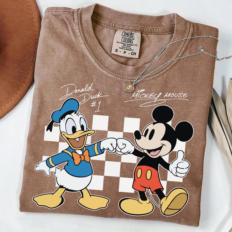May include: Brown t-shirt featuring Donald Duck and Mickey Mouse fist-bumping, with the text "Donald Duck #1" and "Mickey Mouse". The shirt has a checkerboard pattern and a Comfort Colors tag.