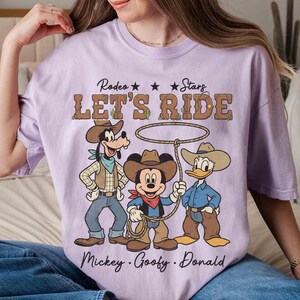 May include: A light purple t-shirt featuring a graphic of Mickey Mouse, Goofy, and Donald Duck dressed as cowboys. The text "Rodeo Stars LET'S RIDE" is above the characters, with their names below.
