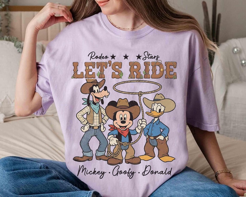 May include: A light purple t-shirt featuring a graphic of Mickey Mouse, Goofy, and Donald Duck dressed as cowboys. The text "Rodeo Stars LET'S RIDE" is above the characters, with their names below.