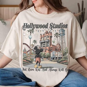 K&ouml;nnte beinhalten: Cremefarbenes T-Shirt mit einer Grafik der Hollywood Studios und Mickey Mouse. Das Design enth&auml;lt den Text "Hollywood Studios" und "That Never Was And Always Will Be". Das Bild zeigt eine Themenpark-Szene mit einer Achterbahn, Palmen und einem Wasserturm.