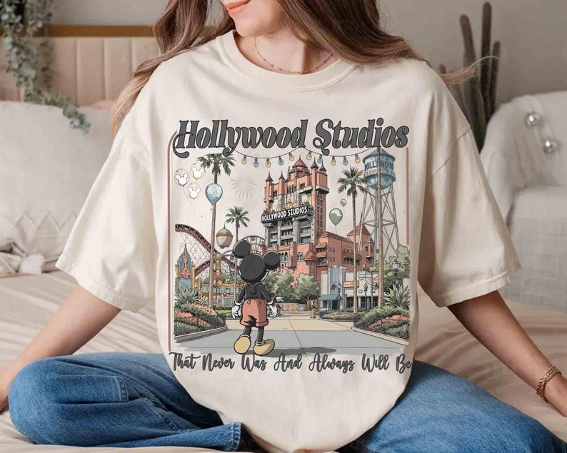 K&ouml;nnte beinhalten: Cremefarbenes T-Shirt mit einer Grafik der Hollywood Studios und Mickey Mouse. Das Design enth&auml;lt den Text "Hollywood Studios" und "That Never Was And Always Will Be". Das Bild zeigt eine Themenpark-Szene mit einer Achterbahn, Palmen und einem Wasserturm.