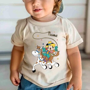 K&ouml;nnte beinhalten: Beiges T-Shirt mit einem Cartoon von Mickey Mouse als Cowboy auf einem Pferd. Das Design beinhaltet einen Seil-Lasso, Sterne und das Wort "Name". Das T-Shirt hat kurze &Auml;rmel.