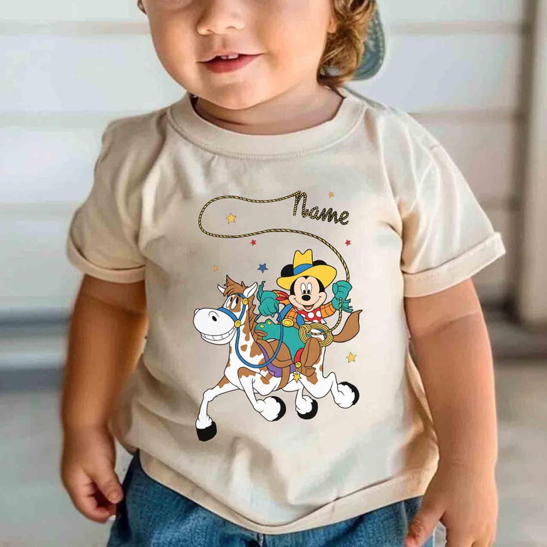 K&ouml;nnte beinhalten: Beiges T-Shirt mit einem Cartoon von Mickey Mouse als Cowboy auf einem Pferd. Das Design beinhaltet einen Seil-Lasso, Sterne und das Wort "Name". Das T-Shirt hat kurze &Auml;rmel.