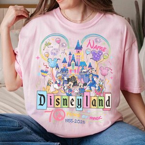May include: Pink t-shirt featuring a colorful Disneyland graphic with Mickey Mouse, Minnie Mouse, Donald Duck, and other Disney characters. The design includes a castle, balloons, and the text "Disneyland 70 Years of Magic 1955-2025".