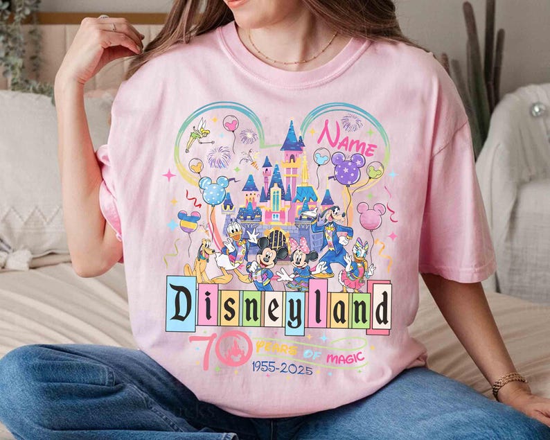 May include: Pink t-shirt featuring a colorful Disneyland graphic with Mickey Mouse, Minnie Mouse, Donald Duck, and other Disney characters. The design includes a castle, balloons, and the text "Disneyland 70 Years of Magic 1955-2025".
